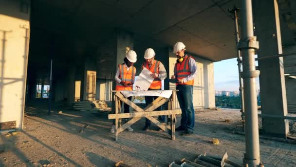 Tres trabajadores de la construcción miran un plano en un sitio de construcción. Ingenieros multiétnicos, arquitectos discutiendo plan de construcción . — Vídeos de Stock