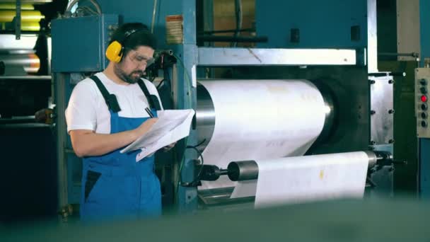 Mannelijke werknemer maakt aantekeningen naast een werkende drukpers — Stockvideo