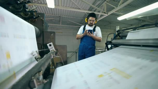 Un trabajador con un smartphone está de pie cerca de una máquina de impresión — Vídeos de Stock