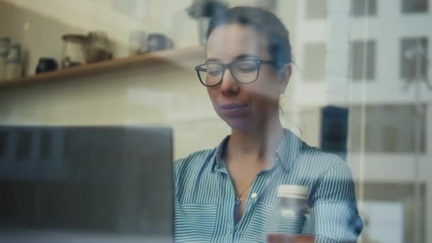 Женщина, работающая на ноутбуке, которую видели через стекло — стоковое видео