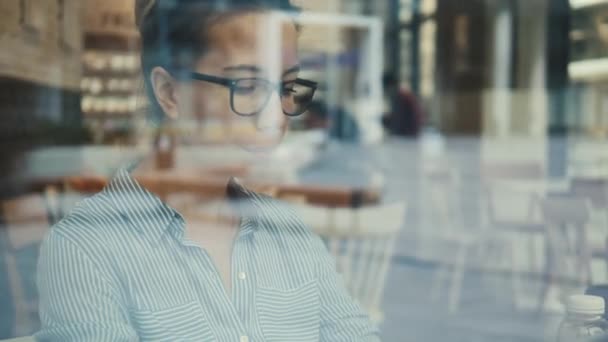 Een vrouw met een bril gefilmd door het raam van het café — Stockvideo