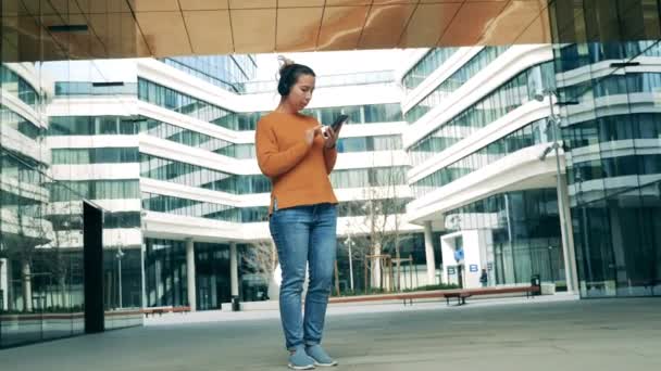 Moderne stad met een dame in een koptelefoon met behulp van haar telefoon — Stockvideo