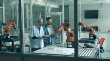 Genç uzmanlar, nesneleri taşıyan robotik bir kolu gözlemliyorlar.