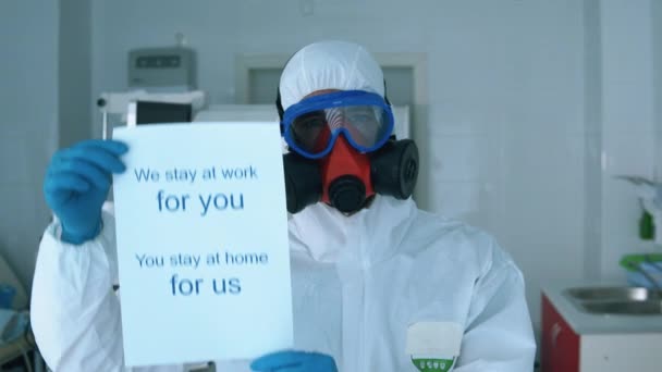 Specialist in een beschermingspak vraagt om thuis te blijven met een poster — Stockvideo