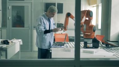 Dizüstü bilgisayarı olan bir erkek bilim adamı hareket halindeki bir robotik cihazı gözlemliyor.