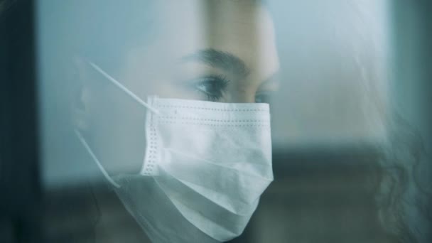 Una donna in maschera medica guarda la finestra mentre rimane a casa . — Video Stock