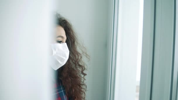 Молодая женщина в маске смотрит в окно во время пандемии коронавируса . — стоковое видео