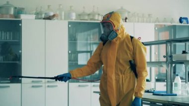 Tehlikeli madde kıyafeti giymiş bir adam laboratuvarı kimyasallarla dezenfekte ediyor.