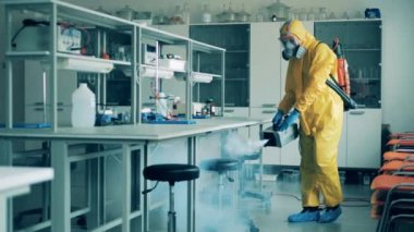 Tehlikeli madde giysisi giymiş bir adam COVID-19 salgını sırasında laboratuvar sınıfını sterilize ediyor..