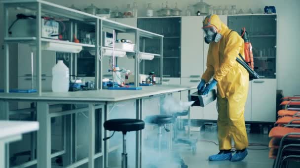 Un hombre en traje de materiales peligrosos está desinfectando un aula de laboratorio durante el brote de COVID-19 . — Vídeos de Stock