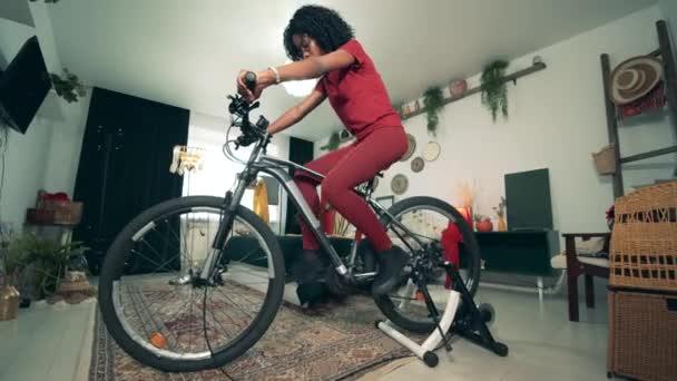 Deporte desde casa, fitness en casa. Mujer afroamericana está montando una bicicleta estática en auto-aislamiento — Vídeo de stock