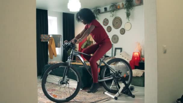 Sport from home, fitness at home. African-American lady is cycling on a fitness machine at home — Stock Video