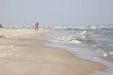Bir kız plaj boyunca bir bisikletin yanına gider denizin arka planına karşı. Uzakta, bir kadın deniz sahilinin temiz havasında yürür..