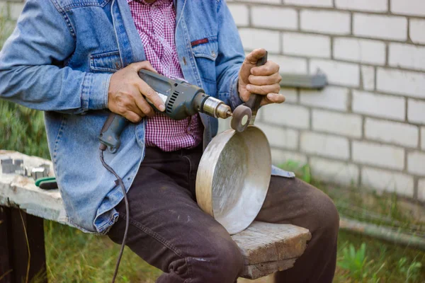 An elderly man cleans the pan mechanically in a personal plot. A man sits with a dirty pan and cleans the burnt fat with a special nozzle.