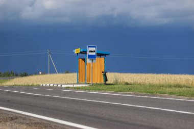Yol kenarındaki köyün yanındaki otobüs durağı Ağustos ayındaki sarı tarla boyunca uzanıyor. Fırtınadan önce olgun bir tarlanın ortasında boş bir yol ve otobüs durağı..
