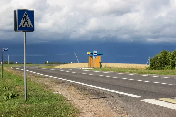 Bus stop near the village on the side of the road running along the August yellow field. An empty road and a bus stop in the middle of a ripe field before a thunderstorm.