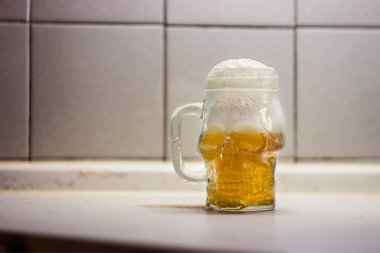 Masanın üzerinde hafif bira olan cam bardak kafatası kavanozu. Bira bardağını beyaz bir zeminde insan kafatası şeklinde tut..