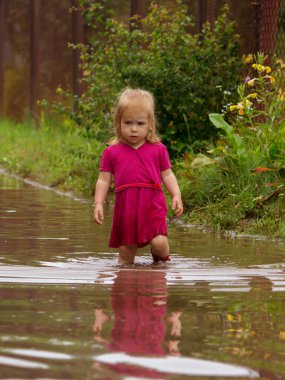 Lastik çizmeli küçük bir kız yağmurdan sonra kumlu bir yolda kirli, derin bir su birikintisinin ortasında duruyor..