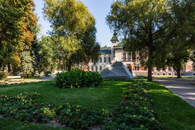 Çiçekli bir çiçek tarhı ve güneşli bir günde parlak çatılı bir mimari binanın önündeki parkta bir anıt. Yatay oryantasyon. Yüksek kalite fotoğraf. Parlak doymuş renkler.