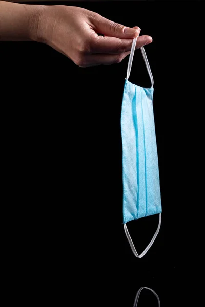 Blue disposable medical mask hanging in the fingers on a black background. Vertical orientation. — Stock Photo, Image