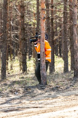 Parlak turuncu ceketli büyük kameralı bir kameraman gülüyor ve ormanda ince bir çam ağacının arkasına saklanıyor. Dikey yönelim.