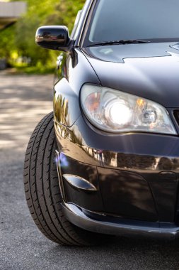 Güneşli bir günde siyah araba parazitli. Arabanın ön tarafı, farlar ve tekerlek. Dikey yönelim.