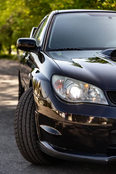 Black car in static on a sunny day. Front view half of the car - headlight and a turned wheel. Vertical orientation. — Stock Photo, Image