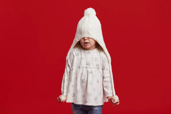 幸せな子供の女の子は彼女の顔にニットの冬の帽子を引く 赤の背景に隔離され — ストック写真