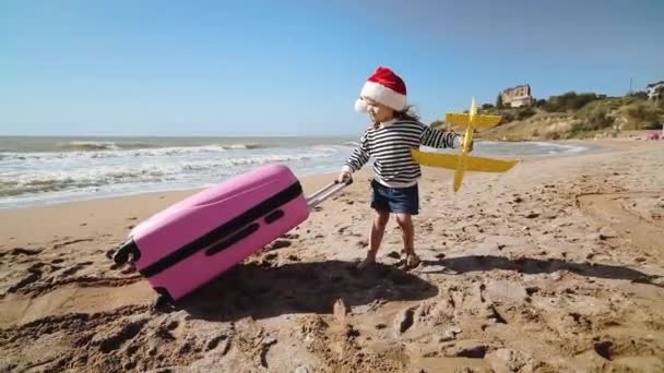 Liten pike i rød juleglue drar koffert i sand – stockvideo