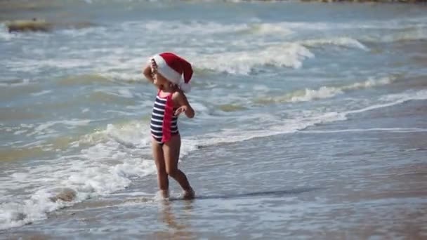 Kırmızı şapkalı, çizgili mayosu olan küçük kız okyanusun kenarında yürüyor. — Stok video