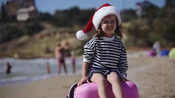 Rolig liten flicka bär röd tomte hatt sitter på resväska — Stockvideo