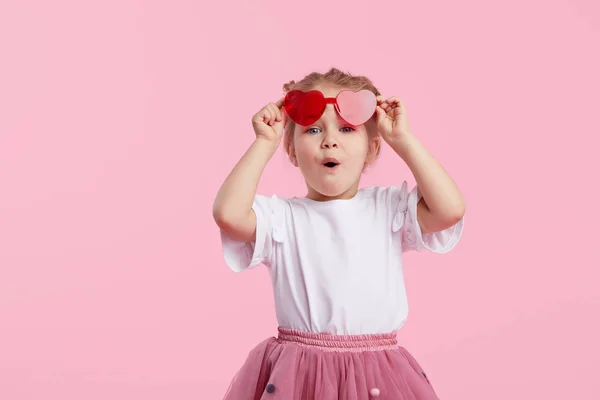 Kalp şeklinde güneş gözlüklü, şaşırmış, küçük kız çocuğunun portresi. Ağzı açık çocuk pembe arka planda izole edilmiş bir şekilde eğleniyor. Kameraya bakıyorum. Komik surat.