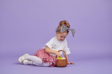 Paskalya tavşanı kostümü giymiş şirin küçük bir kız kocaman bir yumurtanın yanında oturur ve renkli küçük yumurtalarla dolu bir sepet tutar. Mor stüdyo geçmişi. Metin için alanı kopyala