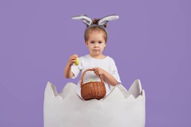 Kostümlü Paskalya tavşanı kulaklı ve yumurtalı sepetli mutlu kız çocuğu. Mor arka planda büyük bir tavuk yumurtası içindeki küçük kız.