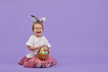 Paskalya tavşanı kostümü giymiş şirin küçük bir kız kocaman bir yumurtanın yanında oturur ve renkli küçük yumurtalarla dolu bir sepet tutar. Mor stüdyo geçmişi. Metin için alanı kopyala