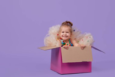 Muhteşem bir tatil elbisesi içinde neşeli küçük bir kız çocuğu mor izole bir arka planda, büyük bir hediye renkli kutunun içinde oturuyor. Doğum günü partisi sürprizi. Metin için boşluk