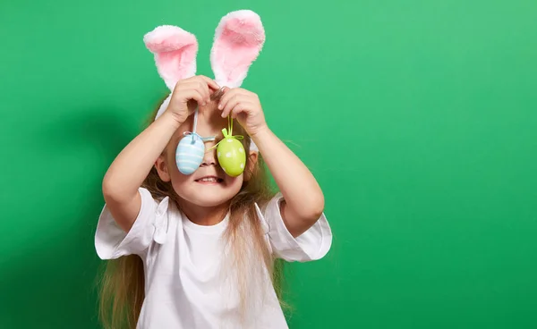 ウサギの耳を持つかわいい小さな子供の女の子と色の緑の背景にイースターエッグ 面白い顔だ — ストック写真