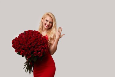 8 Mart ya da Sevgililer Günü hediyesi olarak büyük bir buket kırmızı gül tutan mutlu sarışın kadın. Parmağındaki nişan yüzüğünü gösteriyor. Tarafsız gri stüdyo geçmişi
