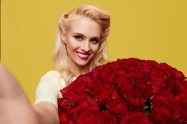 Retro giysili genç bir kadın cep telefonunda parlak sarı arka planda büyük bir buket kırmızı gülle selfie çekiyor. Sevgililer Günü ve 8 Mart için hediye konsepti..