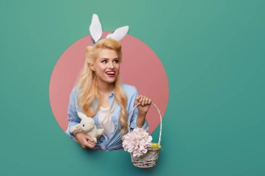 Paskalya tavşanı kulaklı, renkli yumurta sepeti olan genç bir kadın duvardaki yuvarlak delikten sürünerek çıkıyor. Mentollü ya da turkuaz renk arkaplanı. Tatil kartı boş