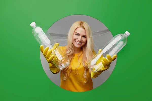 黄色の手袋をした若い女性のボランティアは プラスチックごみの入ったごみ箱が緑の背景に丸い穴を通って見えます 環境保護の概念 — ストック写真