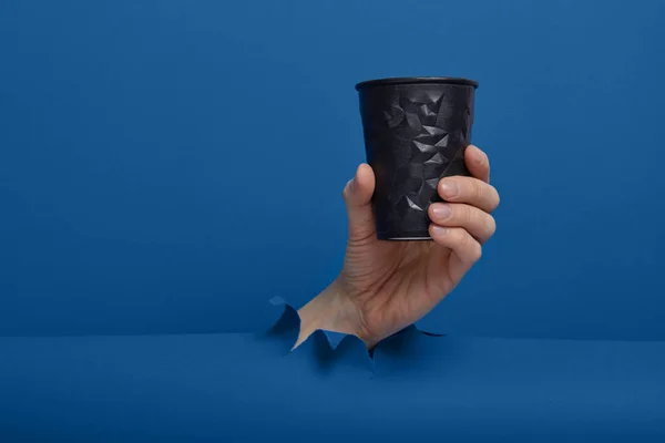 Male hand taking a cup of coffee through torn classic blue paper background. Get energy boost, hot drink in take-away package.