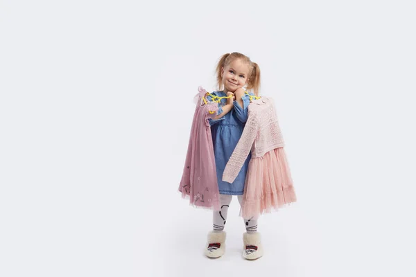 小さい子供の女の子は休日のための服を選ぶ 彼の手にドレスを持つハンガーを保持します ホワイトスタジオの背景 — ストック写真