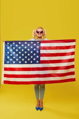 American girl. Happy young woman in denim clothes holding USA flag isolated on yellow background