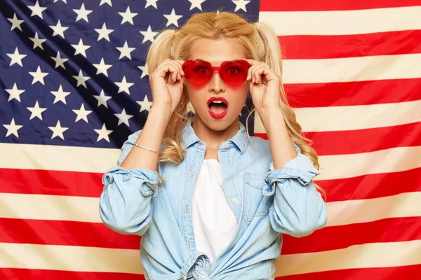 American girl. Happy young woman in heart shape sunglasses on USA flag  background. Funny and surprised human face. Patriotism concept