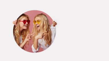 Portrait of two young happy, shocked women in the heart shape sunglasses looking throth the white hole in the wall. Big sale. Funny faces. Empty space for text clipart