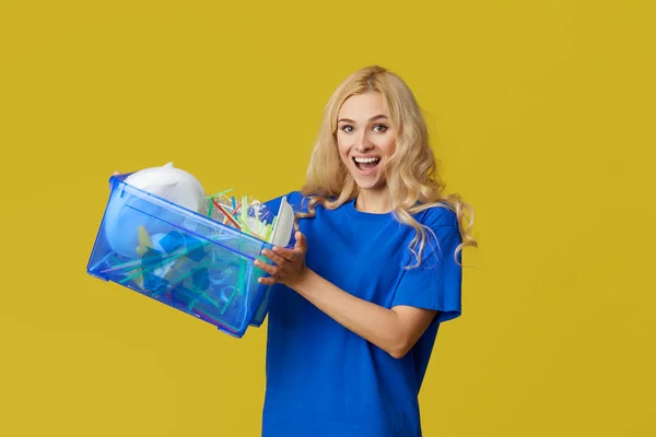 自然への配慮 青いTシャツを着た若い女性がペットボトルや廃棄物の入った箱を持っています ボランティアがゴミをまとめ リサイクルに持ち込みます 黄色の背景 — ストック写真