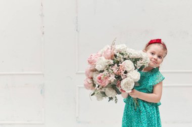 Yazlık elbiseli şirin küçük kız çocuğu beyaz bir duvarın yanında büyük bir buket çiçek tutuyor. Stüdyo cazibesi