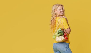 Genç bir kadın sarı arka planda organik sebzelerle dolu bir torba tutuyor. Sıfır atık. Metin için Emty alanı. Sağlıklı Yeme, Vejetaryenlik