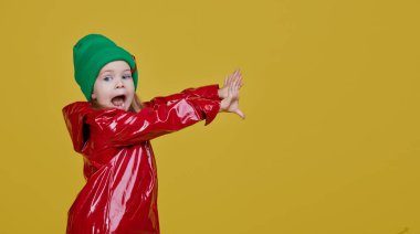 Kırmızı yağmurluk ve sarı arka planda yeşil şapkalı şirin bir kız çocuğu. Komik surat. Parmağını yana çevirir, boşluğu metin için kopyalar